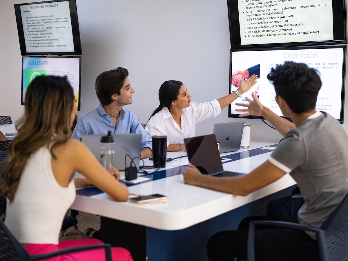 Grupo de estudiantes trabajando en equipo en una sala de clases moderna, con pantallas proyectando contenido académico y laptops sobre la mesa.